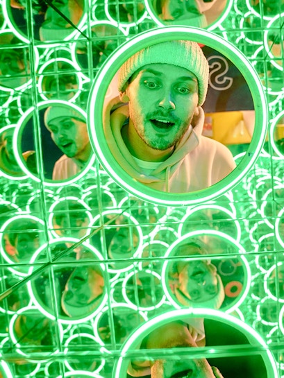 Man in a beanie surrounded by green neon mirrors at World of Illusions, Dublin.