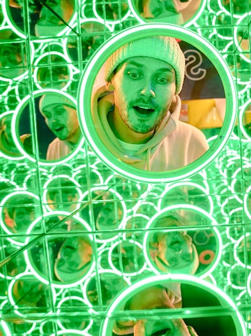 Man in a beanie surrounded by green neon mirrors at World of Illusions, Dublin.