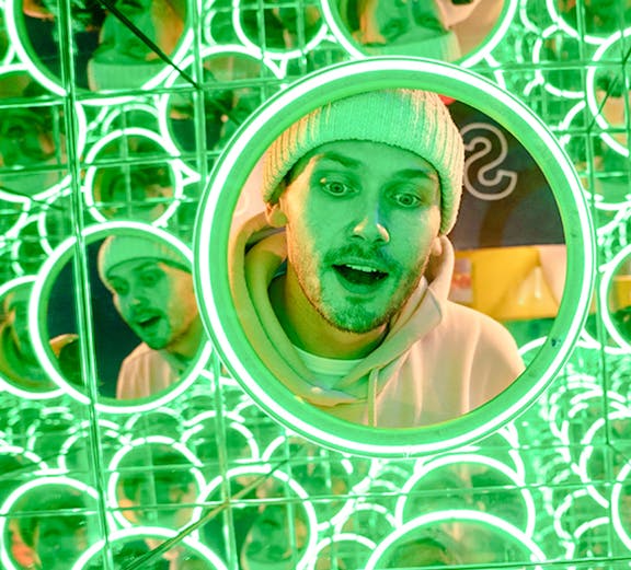 Man in a beanie surrounded by green neon mirrors at World of Illusions, Dublin.
