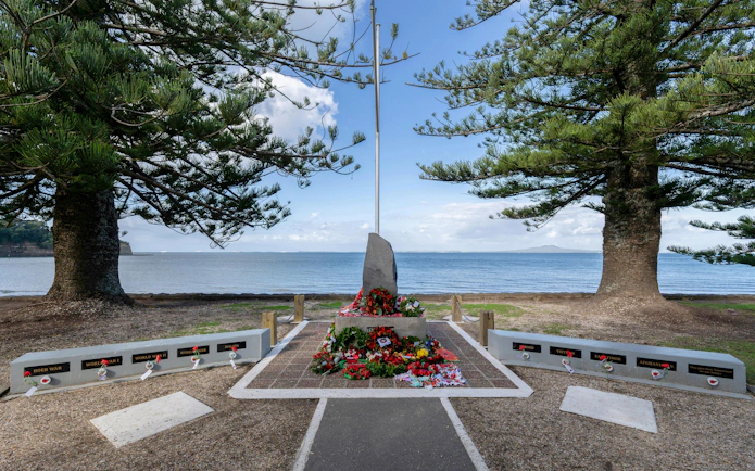 Anzac Memorial with wreaths at Browns Bay Beachfront, New Zealand, between two large trees.