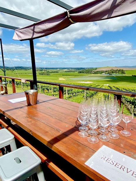 Outdoor seating with wine glasses overlooking Yarra Valley vineyards on a sunny day.