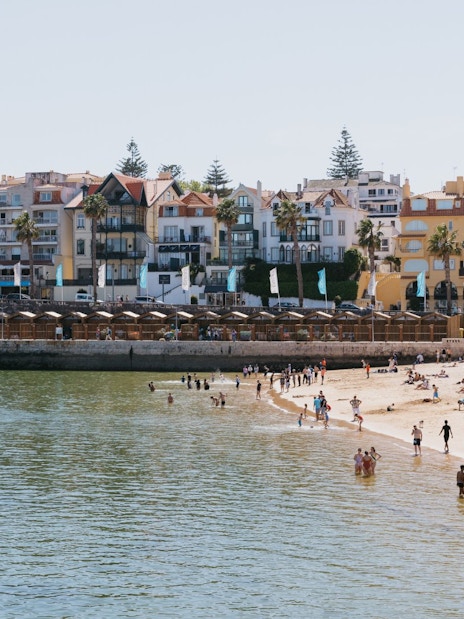 Beachgoers at Praia da Ribeira in Cascais, Portugal, with colorful buildings in the background.