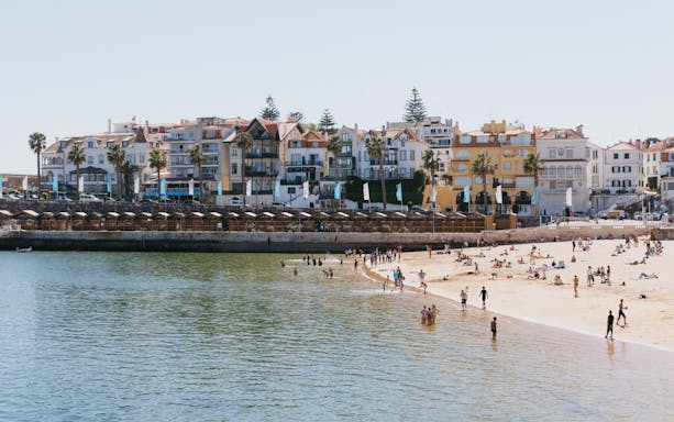 Beachgoers at Praia da Ribeira in Cascais, Portugal, with colorful buildings in the background.