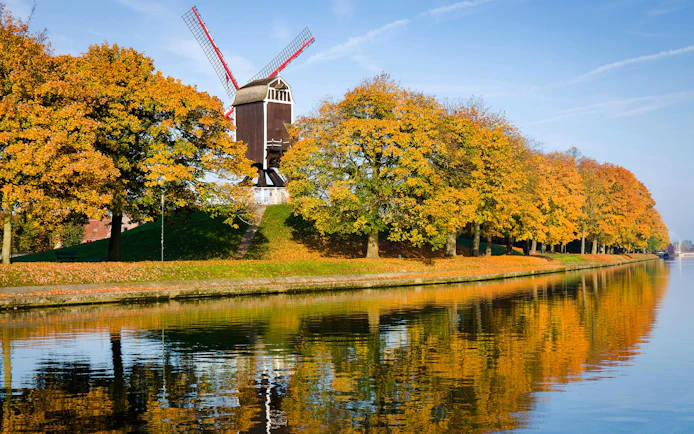 Red wooden windmill at Vesten in Bruges, Belgium, with autumn trees reflected in the water.