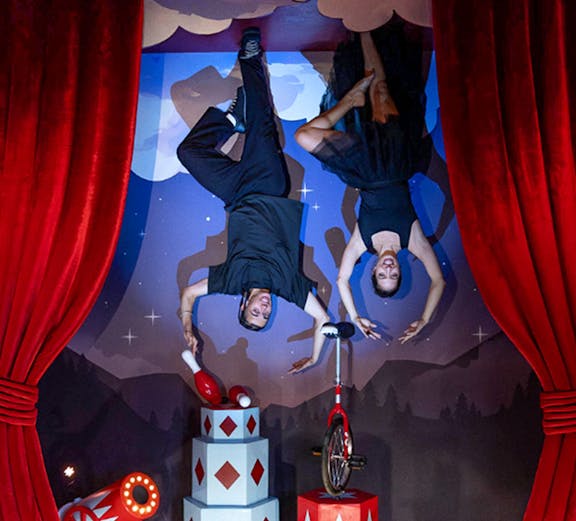 Upside Down Berlin Museum exhibit with performers on ceiling, circus props, and red curtains.