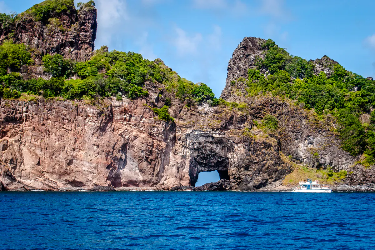 Rocky coastline with lush greenery and a boat near a sea arch, Noronha Boat Tour, Sao Paulo.