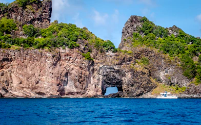 Rocky coastline with lush greenery and a boat near a sea arch, Noronha Boat Tour, Sao Paulo.