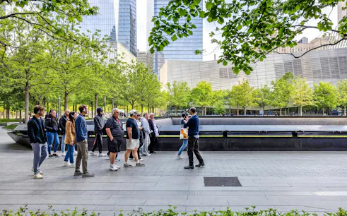 Visitors at the 9/11 Memorial in New York City, with trees and skyscrapers in the background.