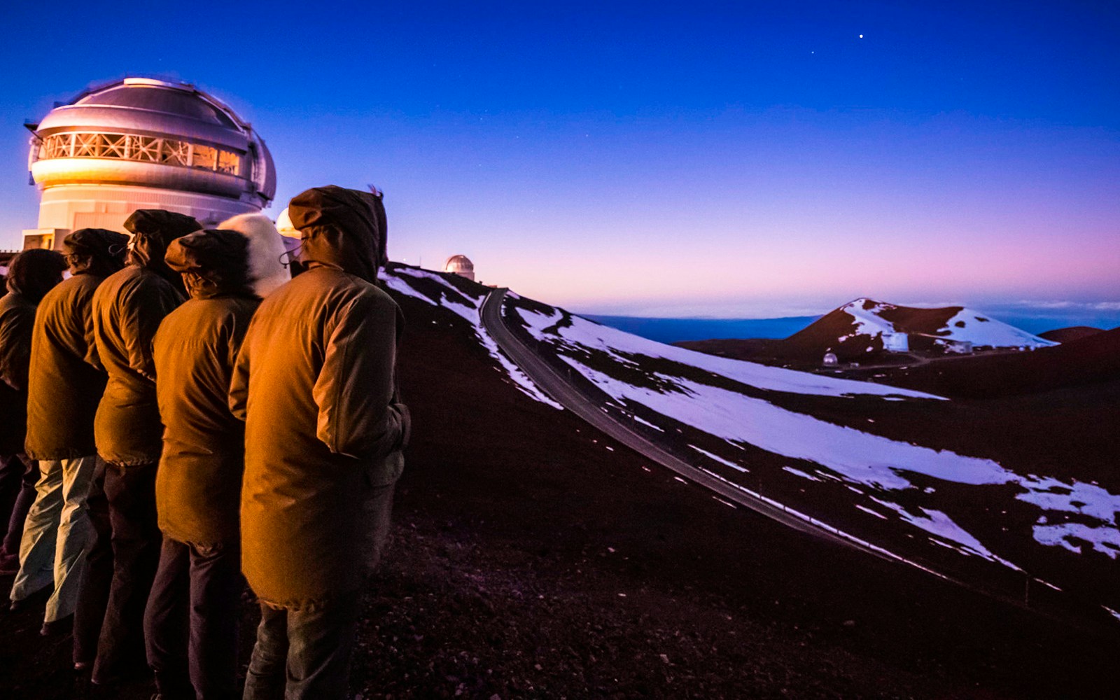 Visitors observing Mauna Kea Observatory at sunset, with snow-covered slopes and a clear sky.