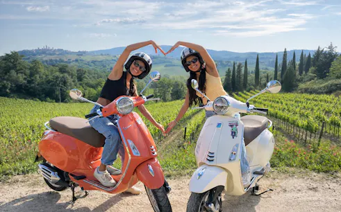 Two people on Vespas making a heart shape with their arms in a Chianti vineyard at sunset.