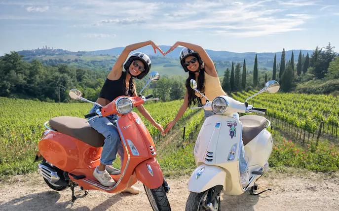 Two people on Vespas making a heart shape with their arms in a Chianti vineyard at sunset.