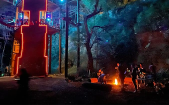 Night zipline tower with people enjoying campfire s'mores in a forest setting.