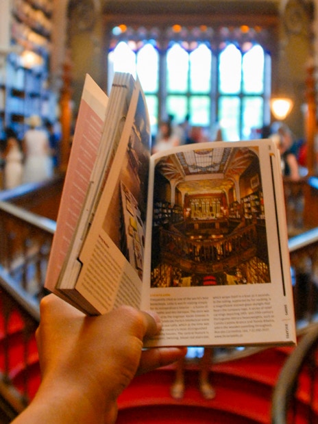 Tourist holding a book inside Lello Library, Porto, surrounded by bookshelves.