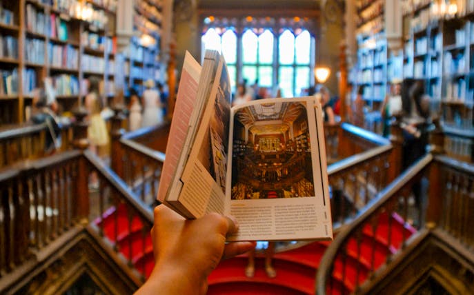 Tourist holding a book inside Lello Library, Porto, surrounded by bookshelves.