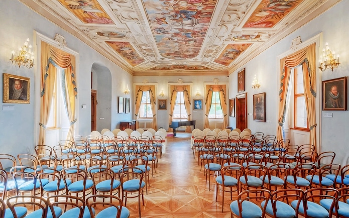 Interior of Lobkowicz Palace concert hall with ornate ceiling and rows of chairs.