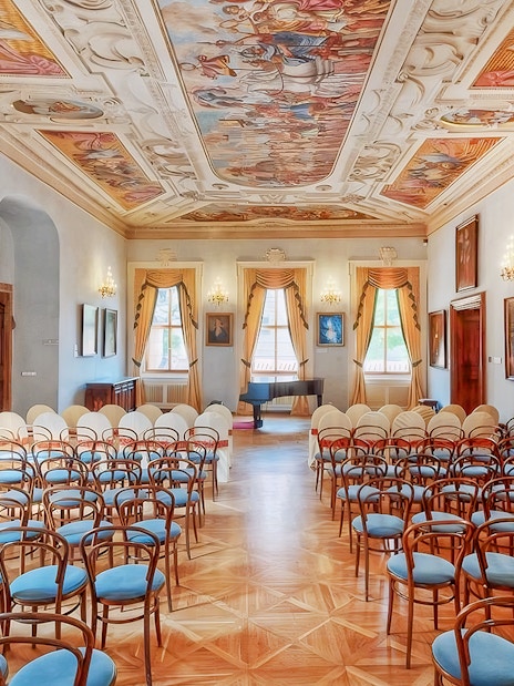 Interior of Lobkowicz Palace concert hall with ornate ceiling and rows of chairs.