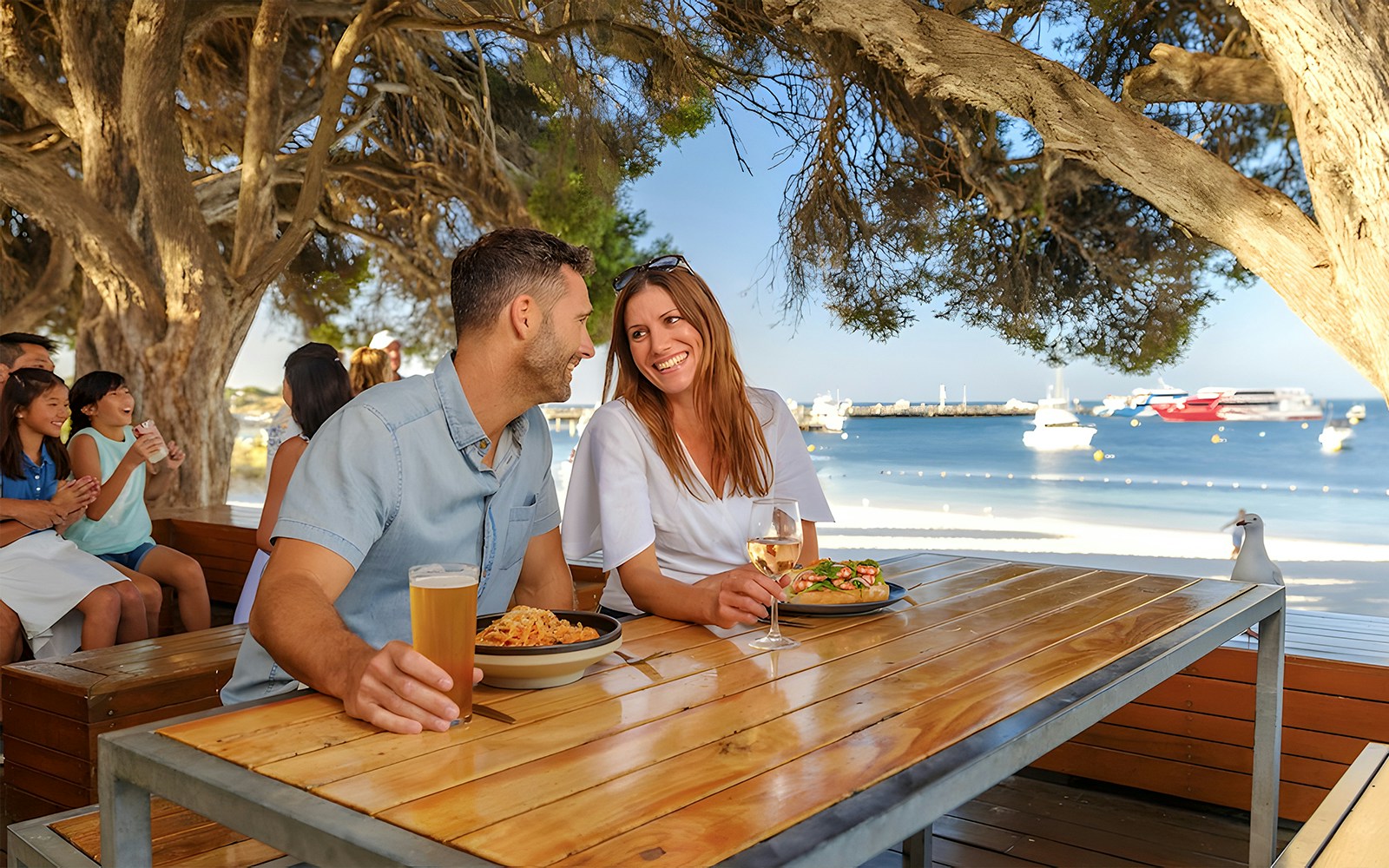 Tourists dining outdoors with a view of Rottnest Island beach and boats in the background.