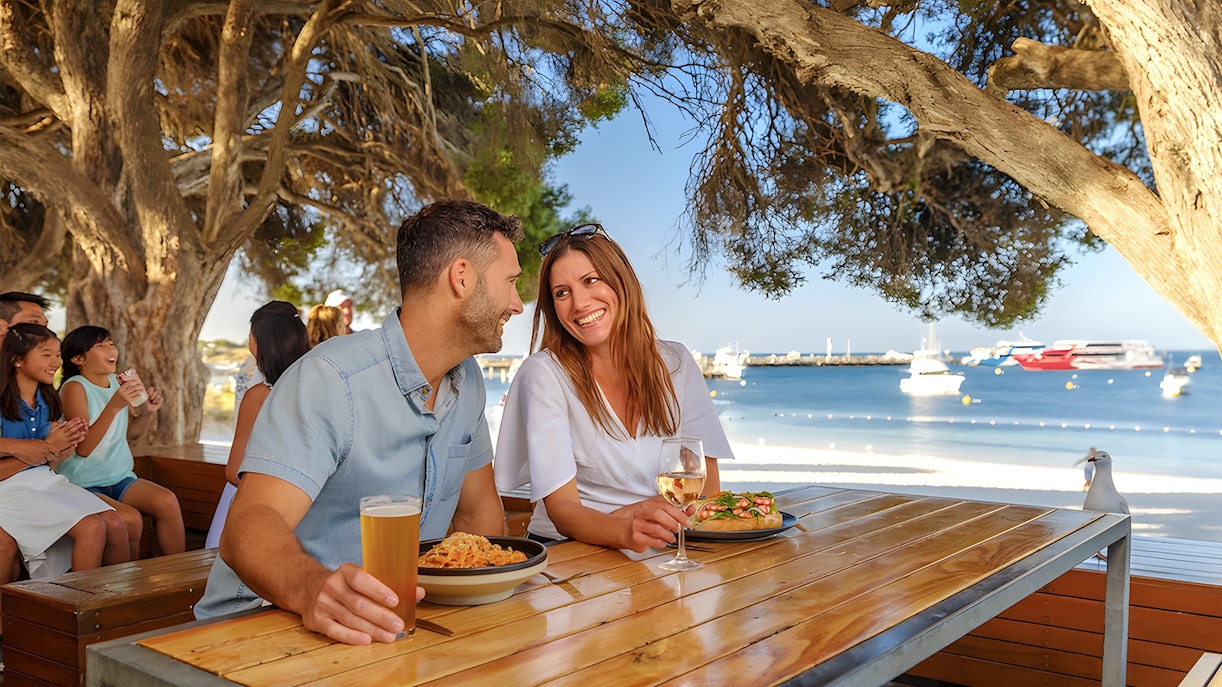 Tourists dining outdoors with a view of Rottnest Island beach and boats in the background.