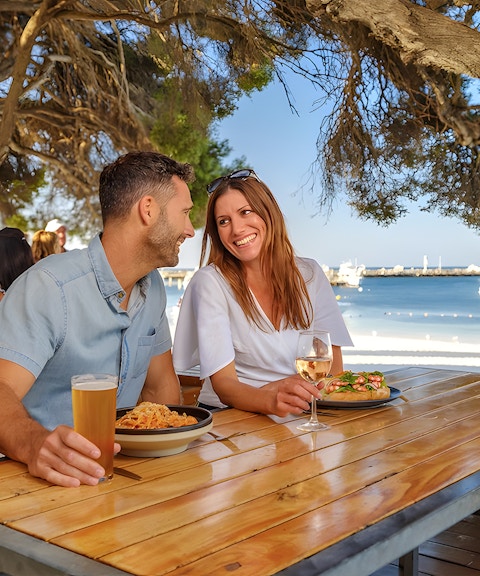 Tourists dining outdoors with a view of Rottnest Island beach and boats in the background.