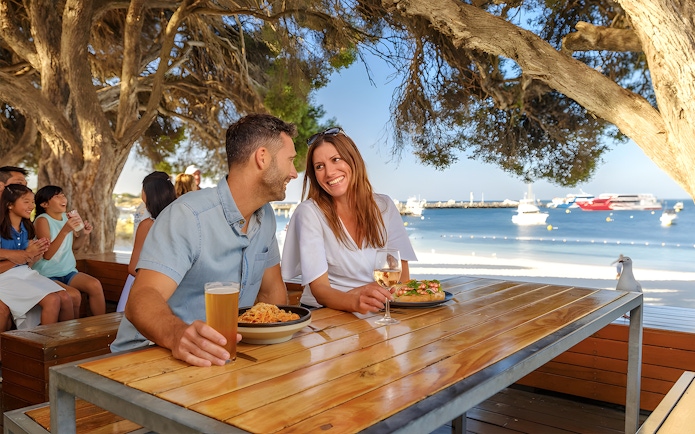 Tourists dining outdoors with a view of Rottnest Island beach and boats in the background.