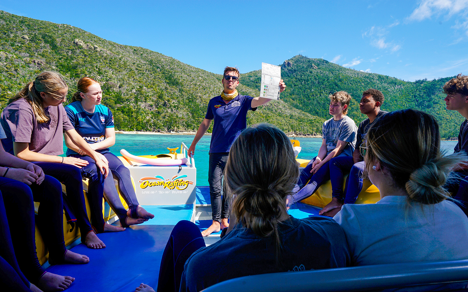 Group on a boat tour near Whitsundays, guide holding a paper, lush hills in the background.