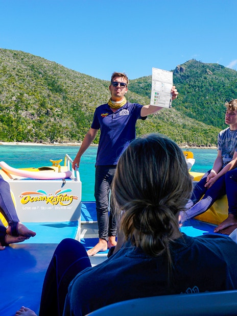 Group on a boat tour near Whitsundays, guide holding a paper, lush hills in the background.
