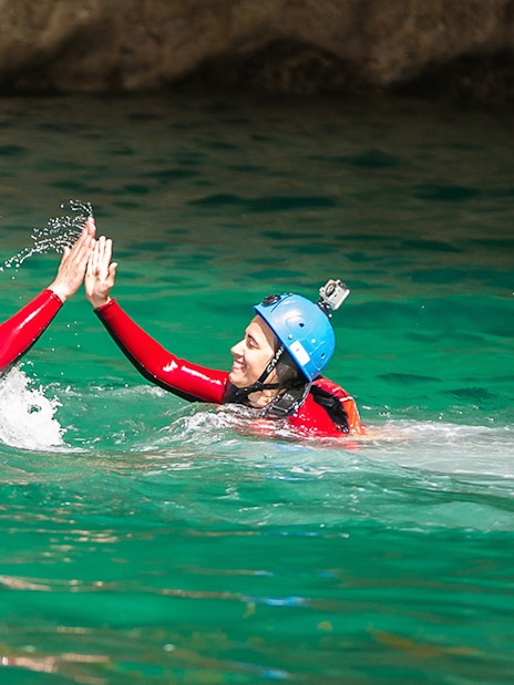 Coasteering participants high-fiving in turquoise water, wearing helmets and wetsuits.