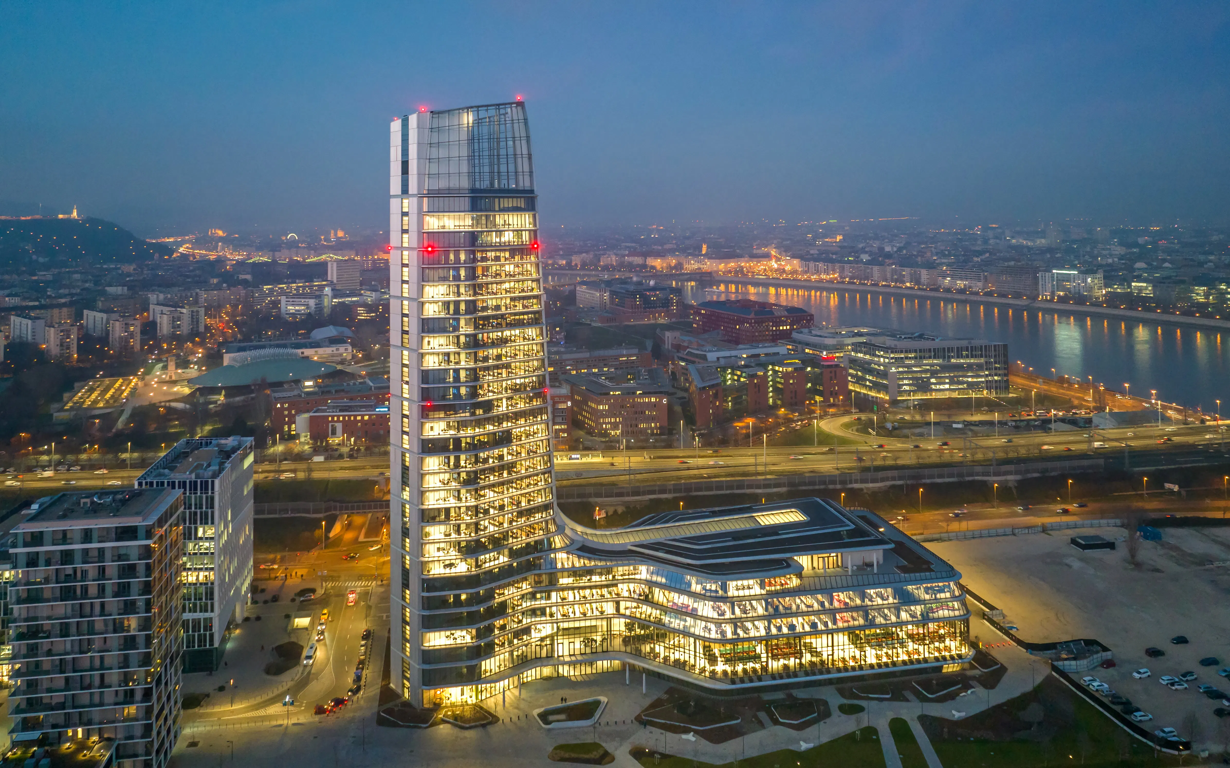 Budapest skyline at night featuring the illuminated MOL HQ skyscraper from a drone view.