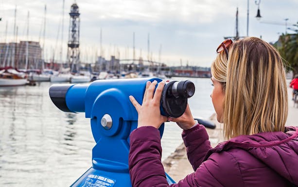 Person using a telescope at Barcelona harbor with boats and cable car tower in view.