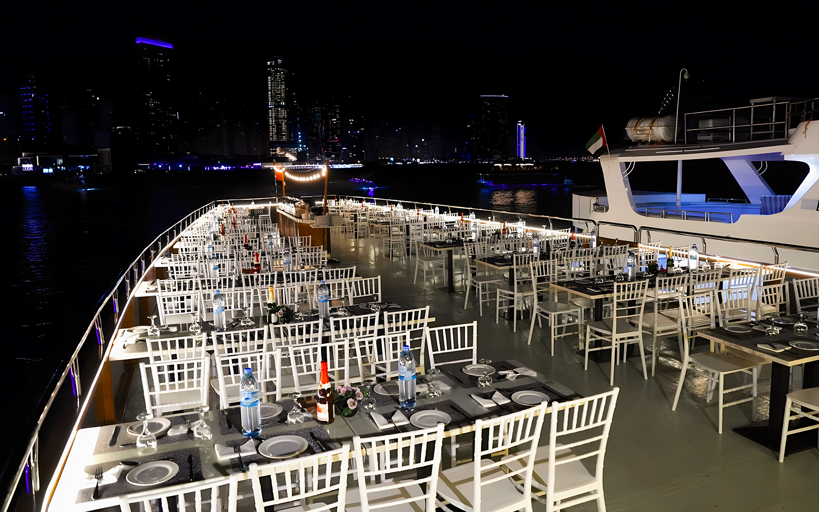 Dhow cruise dinner setup with tables and chairs on deck, Dubai Marina skyline at night.