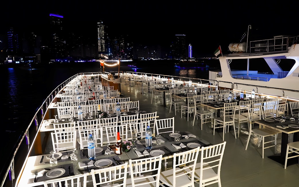 Dhow cruise dinner setup with tables and chairs on deck, Dubai Marina skyline at night.