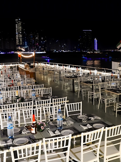 Dhow cruise dinner setup with tables and chairs on deck, Dubai Marina skyline at night.