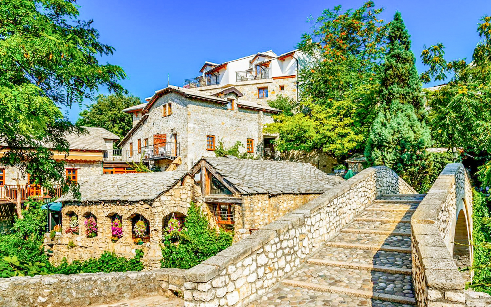 Old stone bridge leading to historic buildings in Mostar, surrounded by lush greenery.