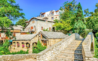Old stone bridge leading to historic buildings in Mostar, surrounded by lush greenery.