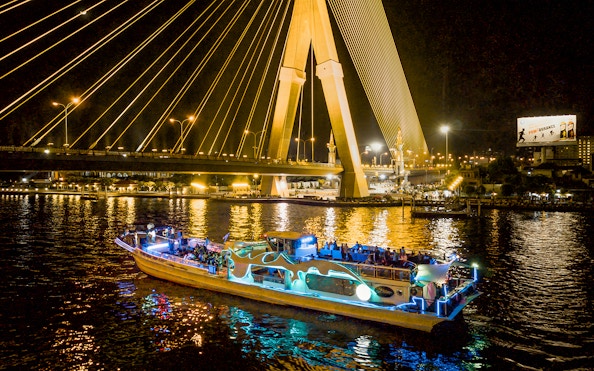 Yodsiam Sightseeing Boat at night under illuminated bridge in Bangkok.