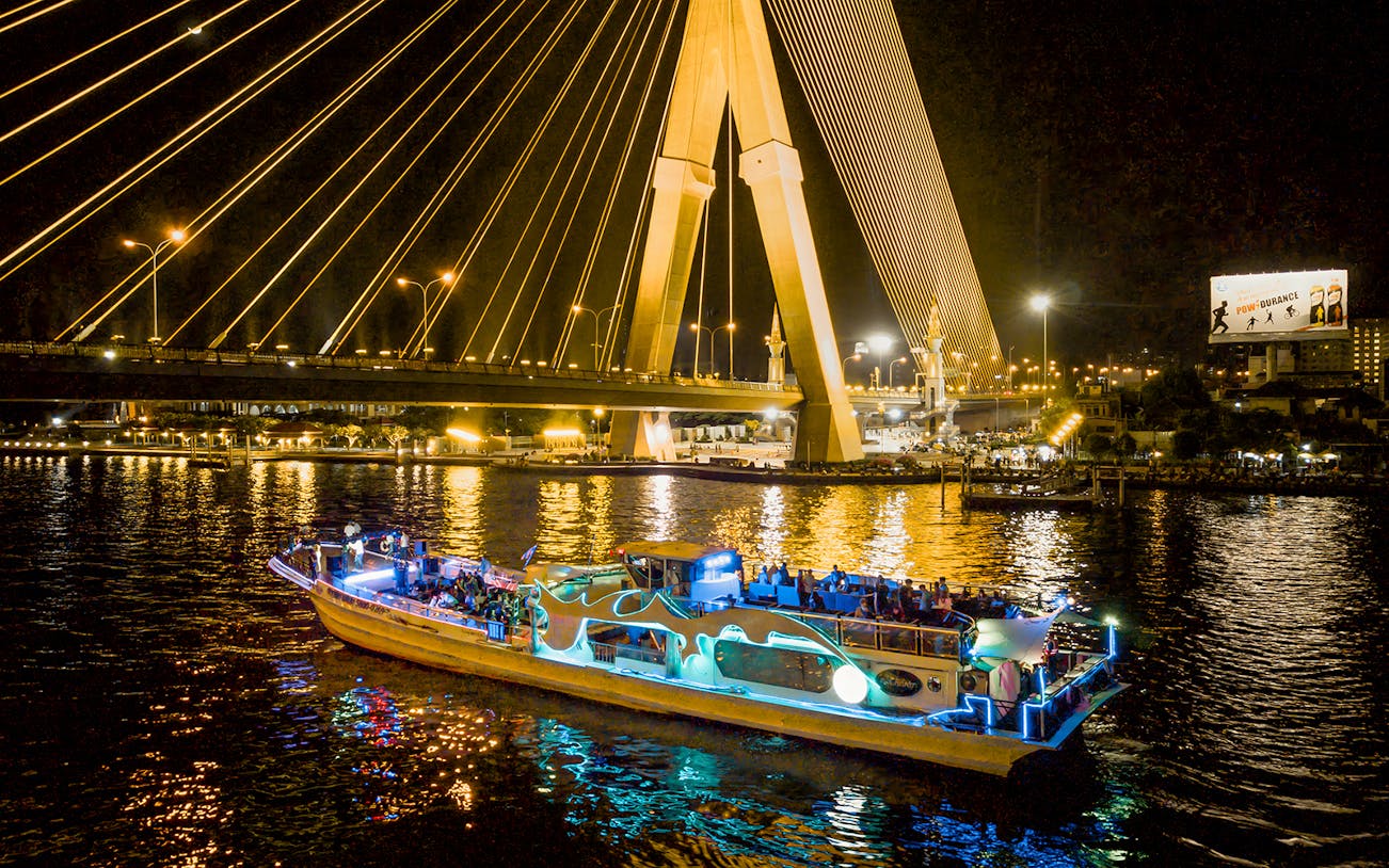 Yodsiam Sightseeing Boat at night under illuminated bridge in Bangkok.