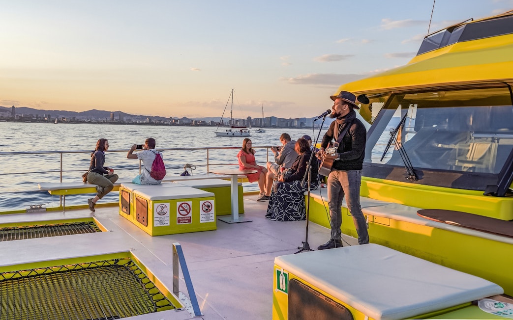 Musician performing on a catamaran cruise at sunset with city skyline in the background.