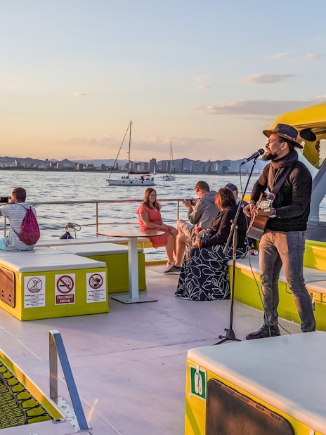 Musician performing on a catamaran cruise at sunset with city skyline in the background.