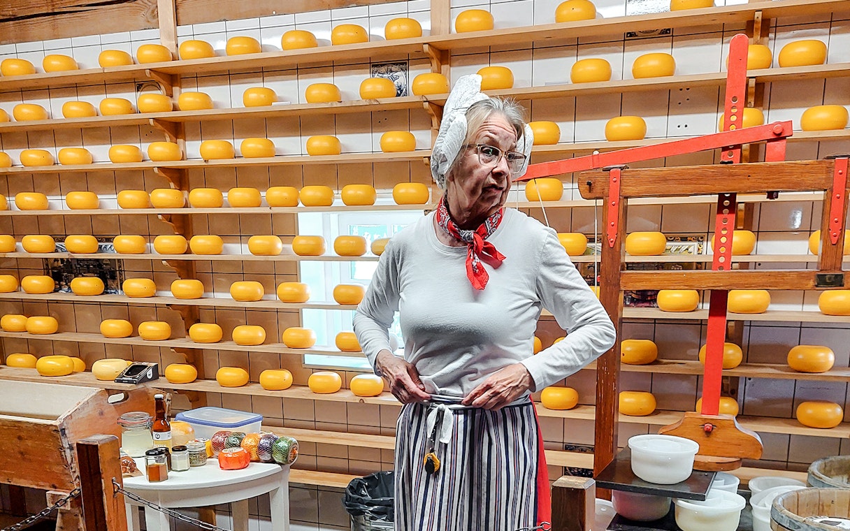 Cheese maker demonstrating process in front of shelves with cheese wheels.