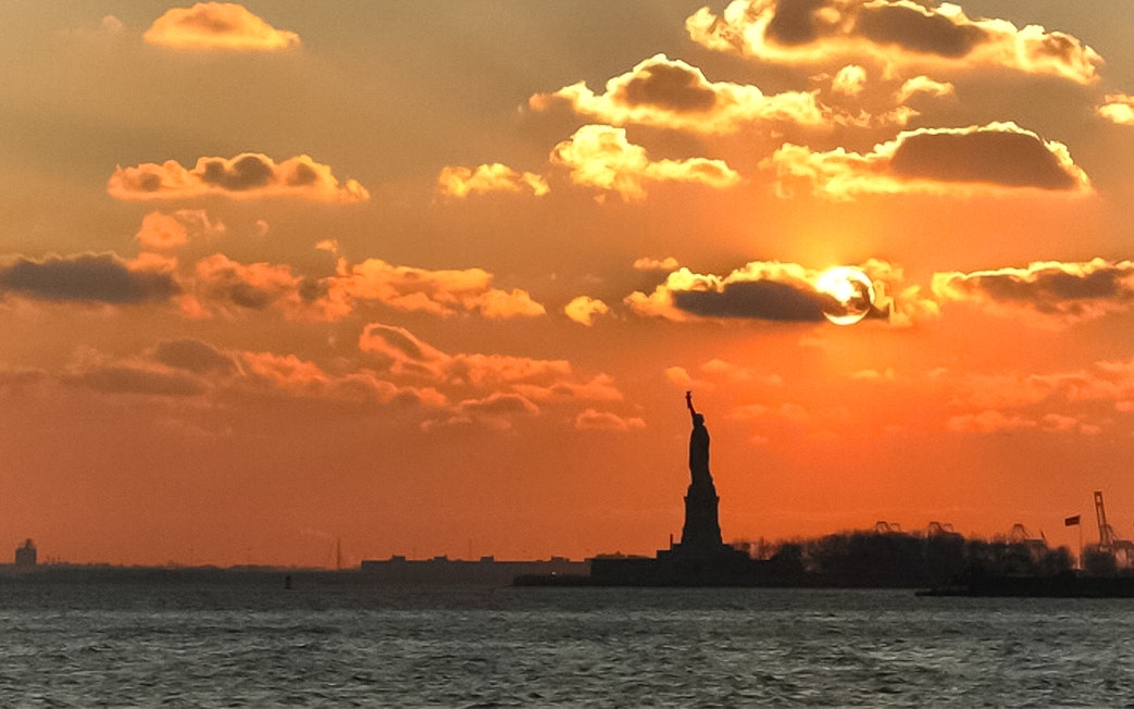 Statue of Liberty silhouetted against sunset sky during Ellis Island cruise.