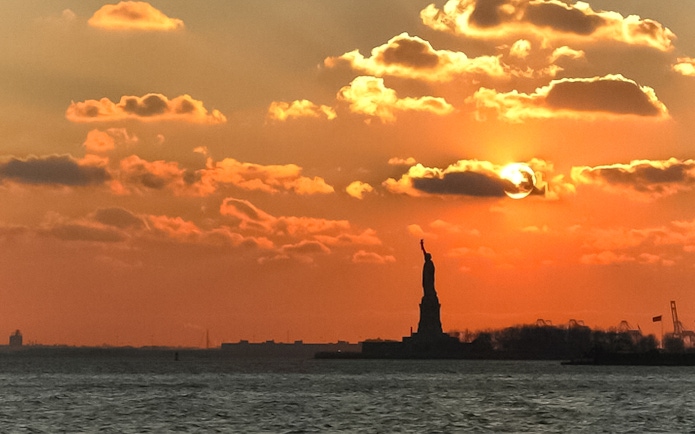 Statue of Liberty silhouetted against sunset sky during Ellis Island cruise.