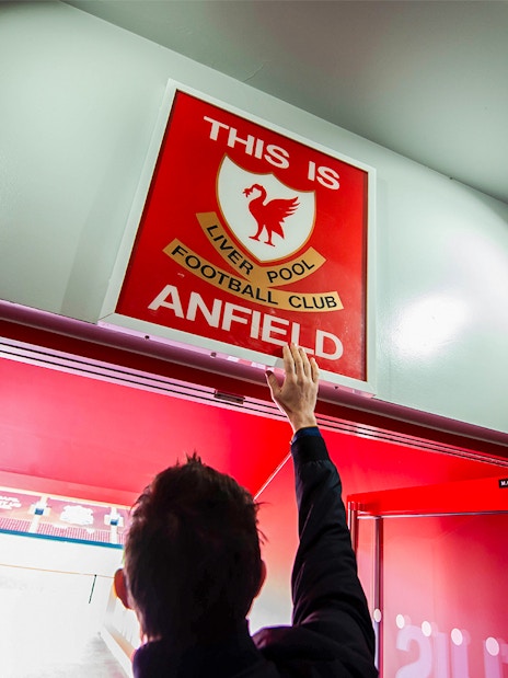Hand reaching for "This is Anfield" sign at Liverpool FC Stadium entrance.
