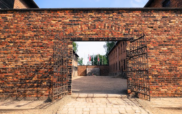 Entrance gate to Auschwitz-Birkenau with brick buildings and flags in the background.