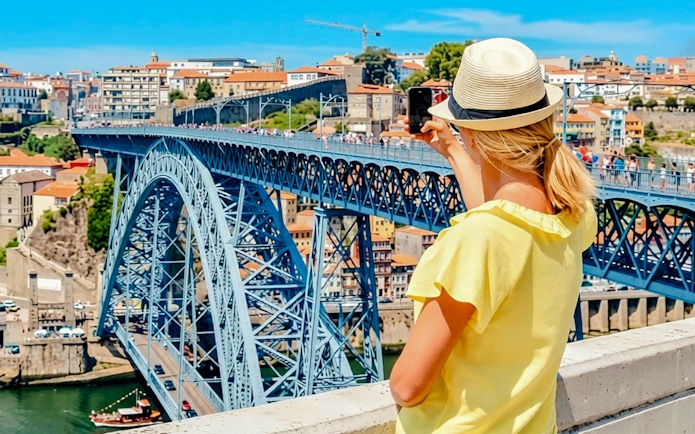 Person taking photo of Dom Luís I Bridge in Porto, with cityscape in the background.