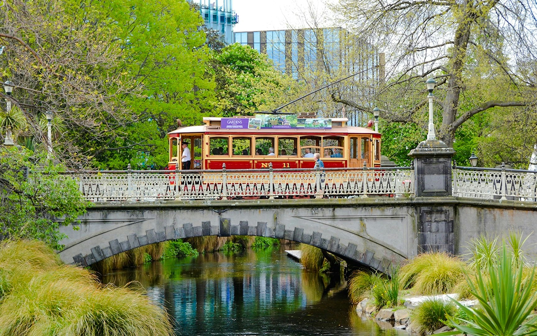 Tram crossing a historic bridge in Christchurch surrounded by lush greenery.