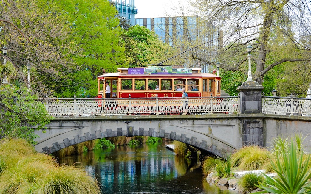 Tram crossing a historic bridge in Christchurch surrounded by lush greenery.
