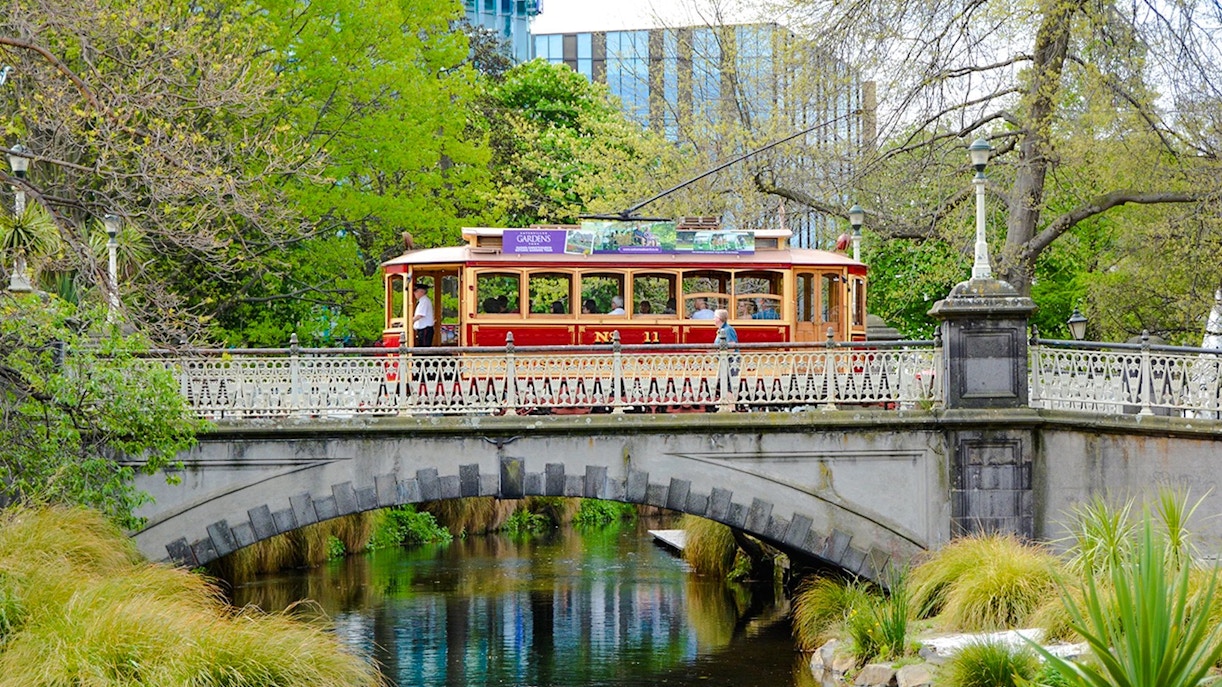 Tram crossing a historic bridge in Christchurch surrounded by lush greenery.
