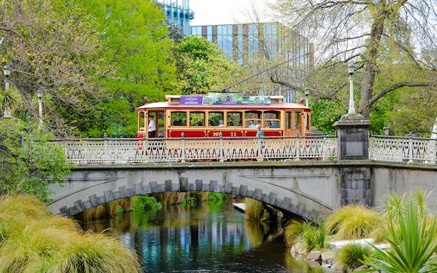 Tram crossing a historic bridge in Christchurch surrounded by lush greenery.