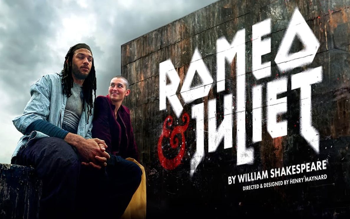Actors in modern attire sit beneath a "Romeo & Juliet" sign, cloudy sky backdrop.