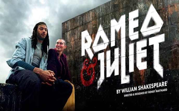 Actors in modern attire sit beneath a "Romeo & Juliet" sign, cloudy sky backdrop.